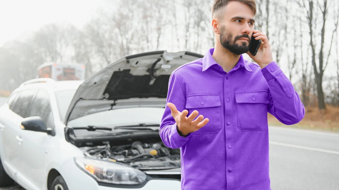 Homem com camisa roxa ao telefone próximo a carro quebrado